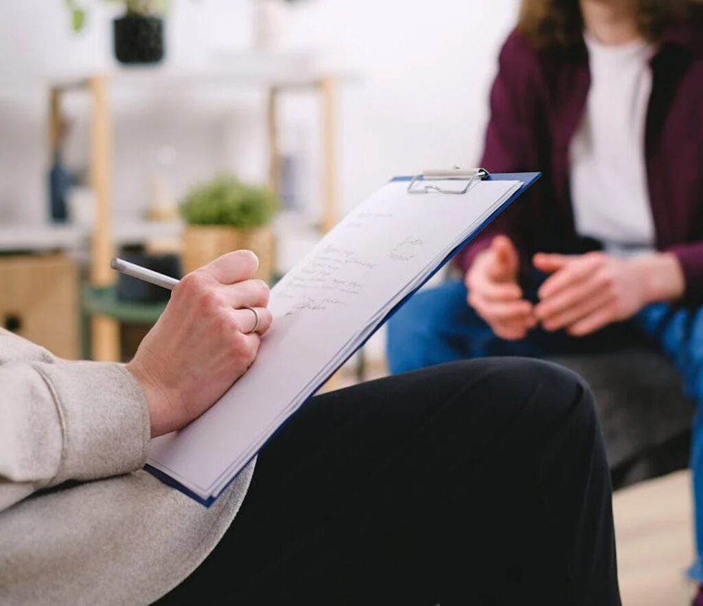 На фото зображена рука, яка щось пише на аркуші паперу. На фоні розмито видно людину, яка сидить навпроти.