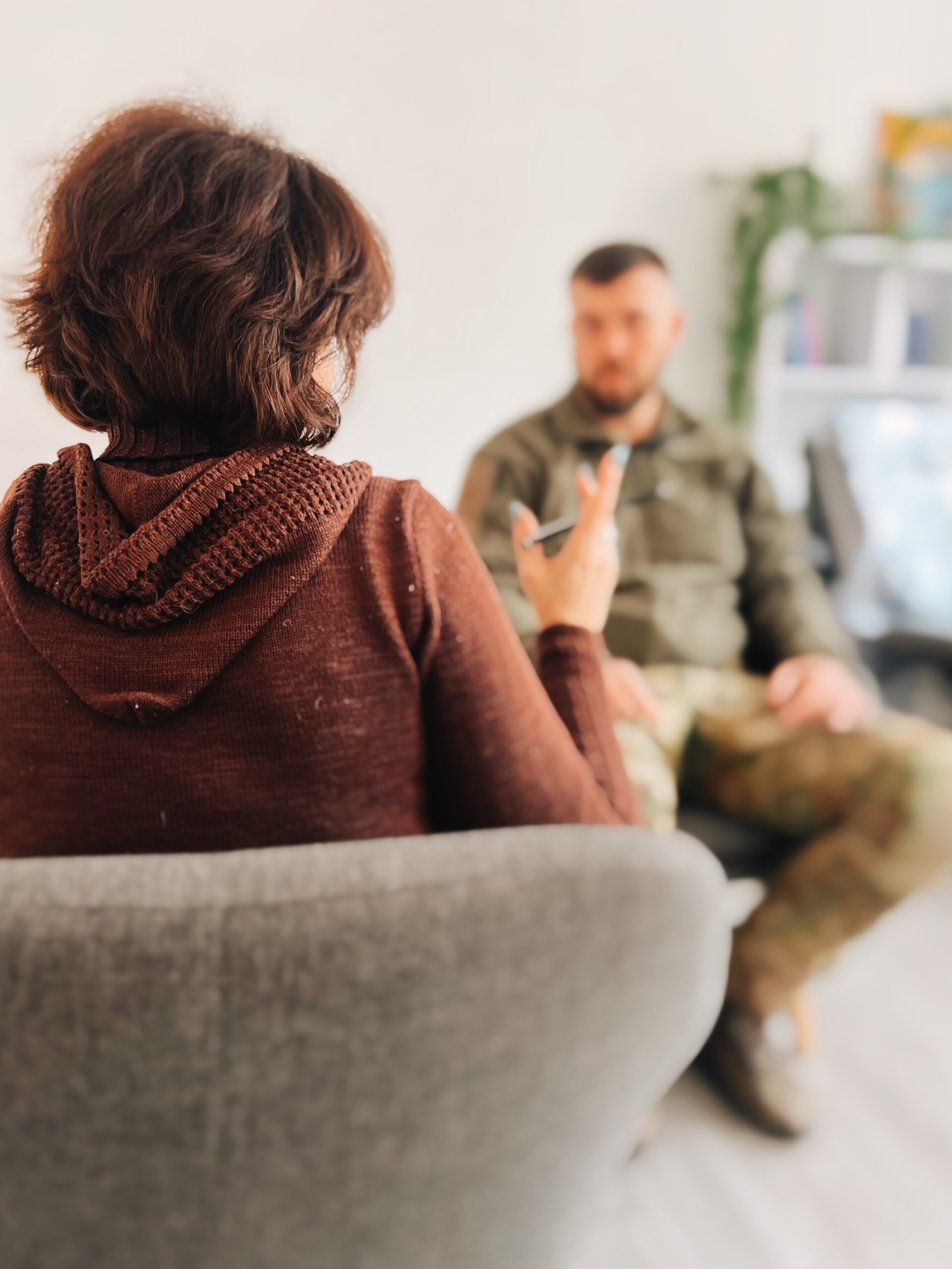 На фото ми зі спини бачимо жінку у кріслі з короткою стрижкою. Вона тримає у руці ручку та щось говорить. Перед нею зображення розмито, але ми можемо побачити силует чоловіка у військовій формі, який також сидить у кріслі та слухає цю жінку.