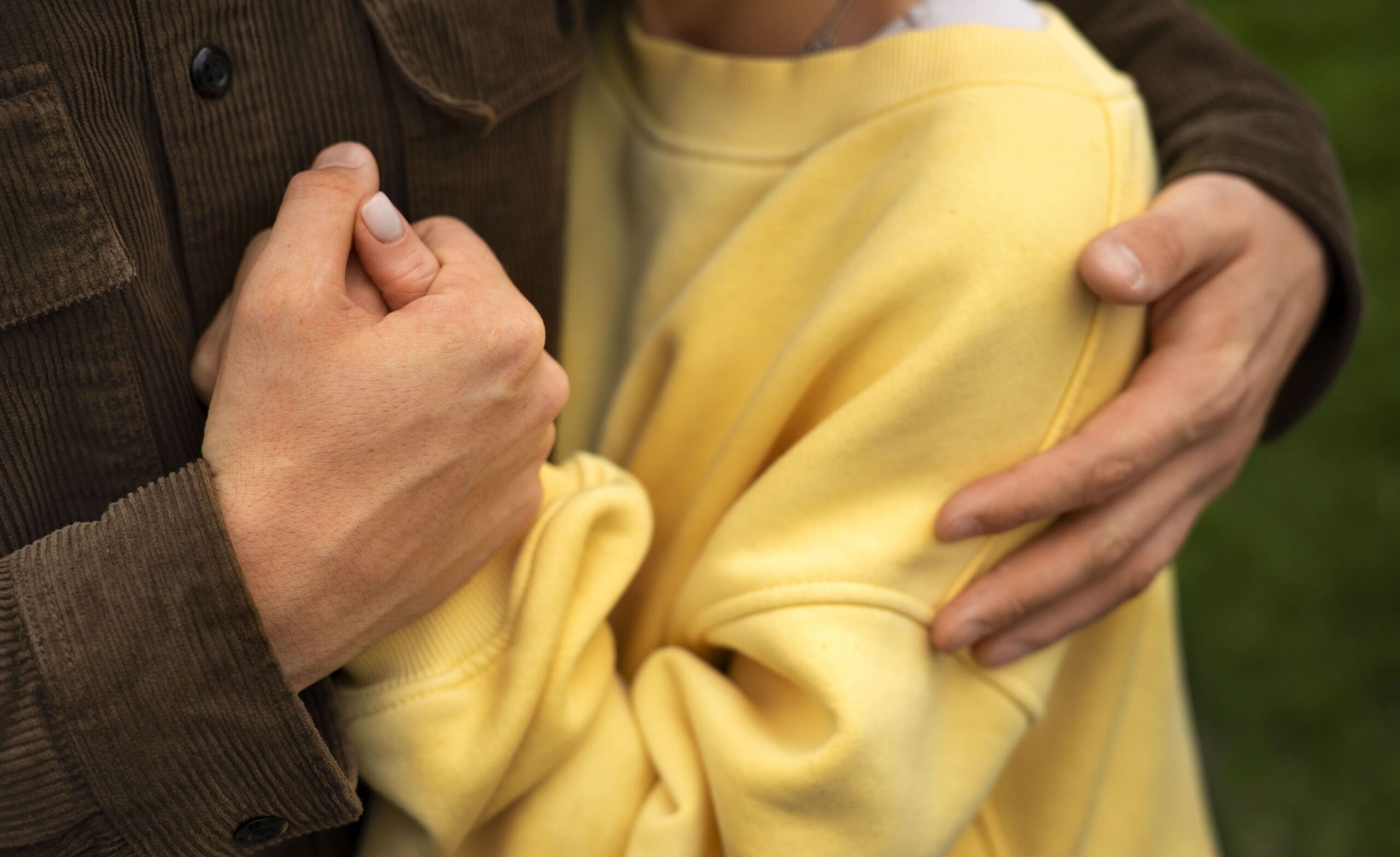 На фото - чоловіча рука тримає жіночу та обіймає жінку за плече. Жінка у жовтому светрі, а чоловік у сорочці кольору хакі.