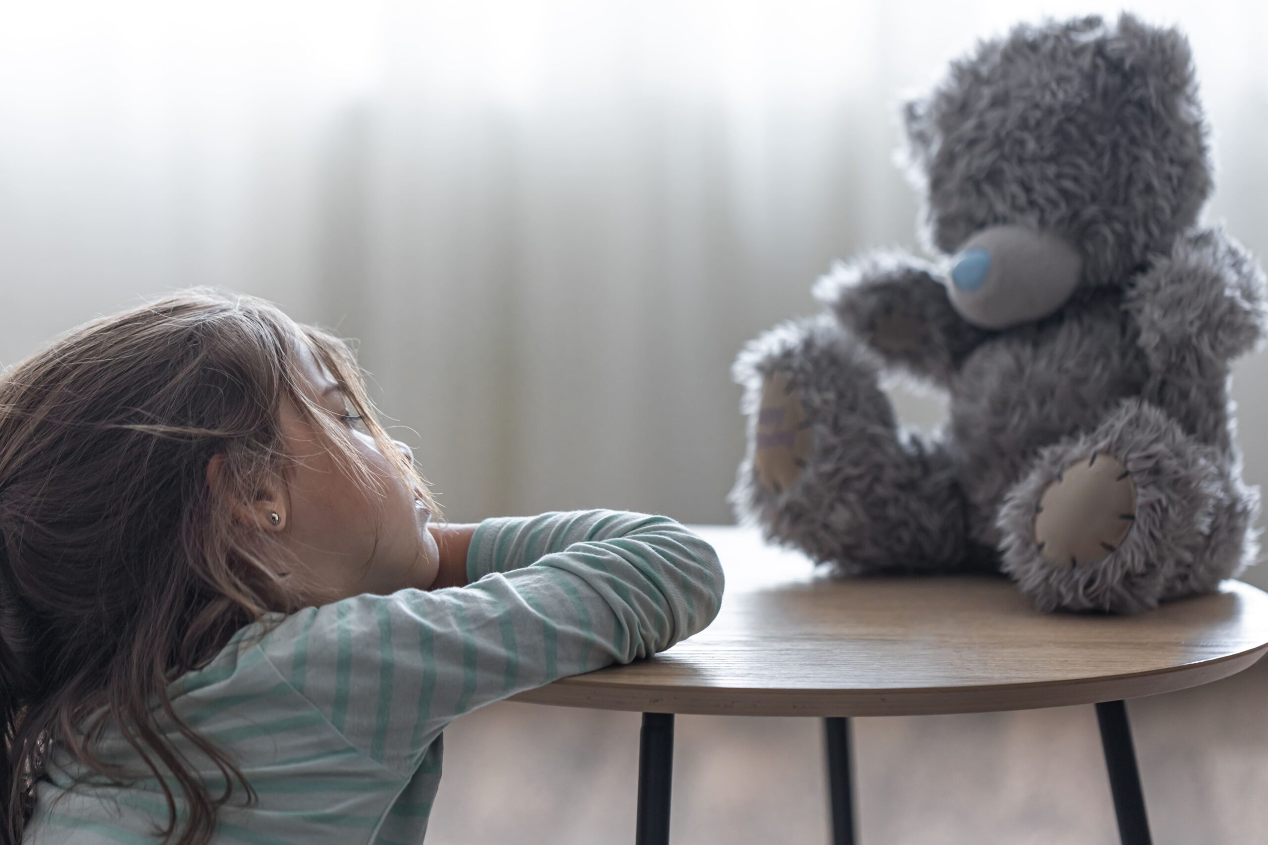 На фото дівчинка сидить на підлозі. Її руки на столі, а голова спирається на них. Дівчинка дивиться на сірого ведмедика, який сидить на столі перед нею. Ймовірно, на її обличчі відображено сум.