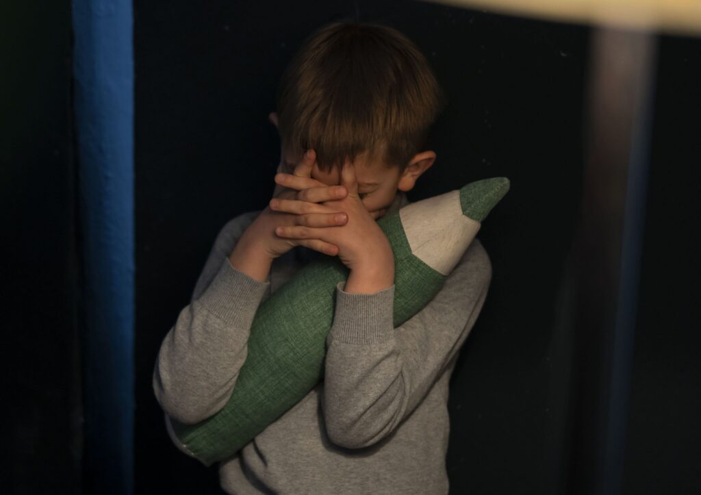 На фото бачимо хлопчика у сірому светрі. Він тримає у руках м'яку іграшку у вигляді зеленого олівця та закриває руками обличчя. Фон навколо темний.