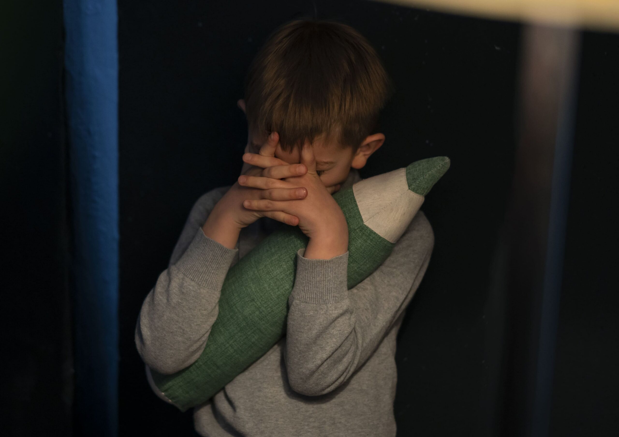 На фото бачимо хлопчика у сірому светрі. Він тримає у руках м'яку іграшку у вигляді зеленого олівця та закриває руками обличчя. Фон навколо темний.