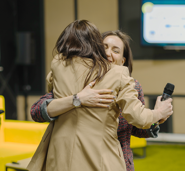 На фото зображені дві жінки, які обіймаються. Одна із них тримає у руках мікрофон, вона одягненна у щось, схоже на піджак бежевого кольору.