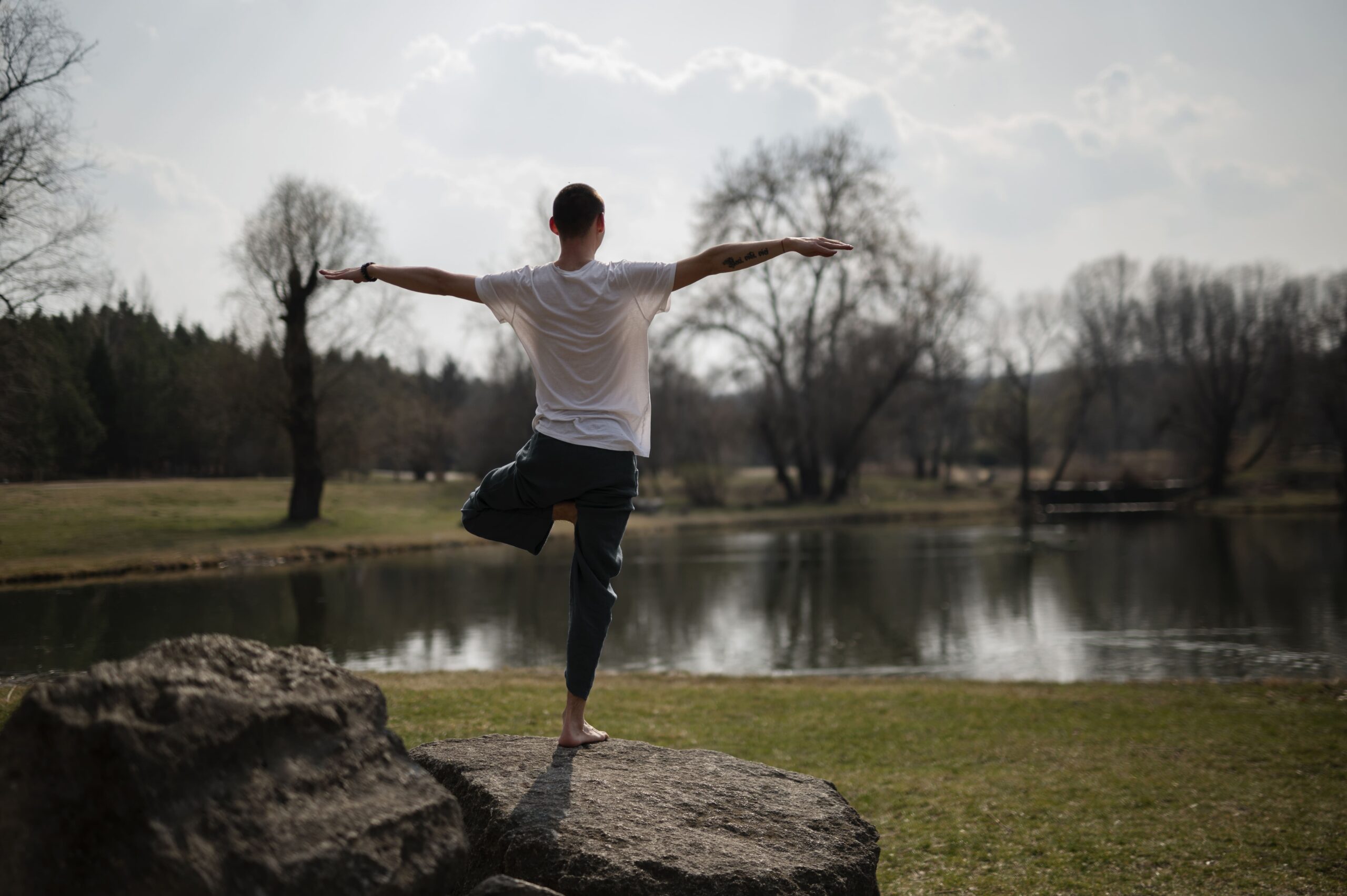 На фото зображений чоловік у повний зріст. Він стоїть до нас спиною на фоні озера, дерев та трави. Чоловік широко розкинув руки, а одну ногу зігнув у коліні, стоячи в одній із поз йоги.