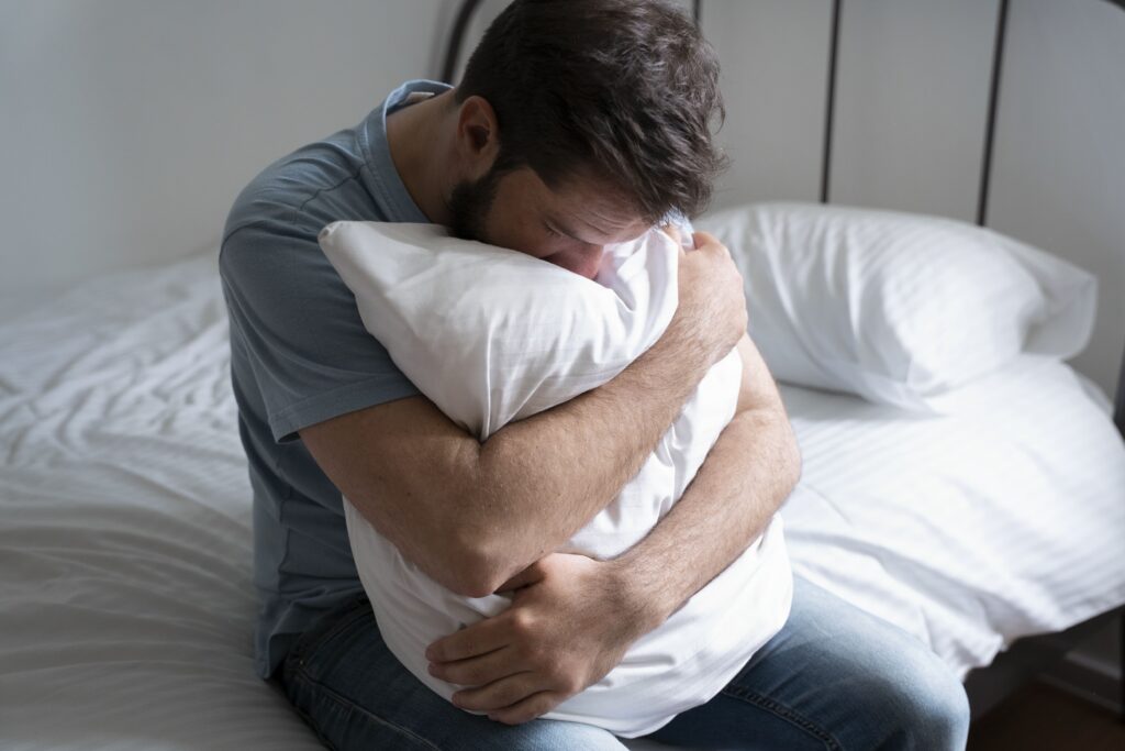 На фото зображений чоловік із темним волоссям та у сірій футболці. Він сидить на ліжку та обіймає обома руками подушку