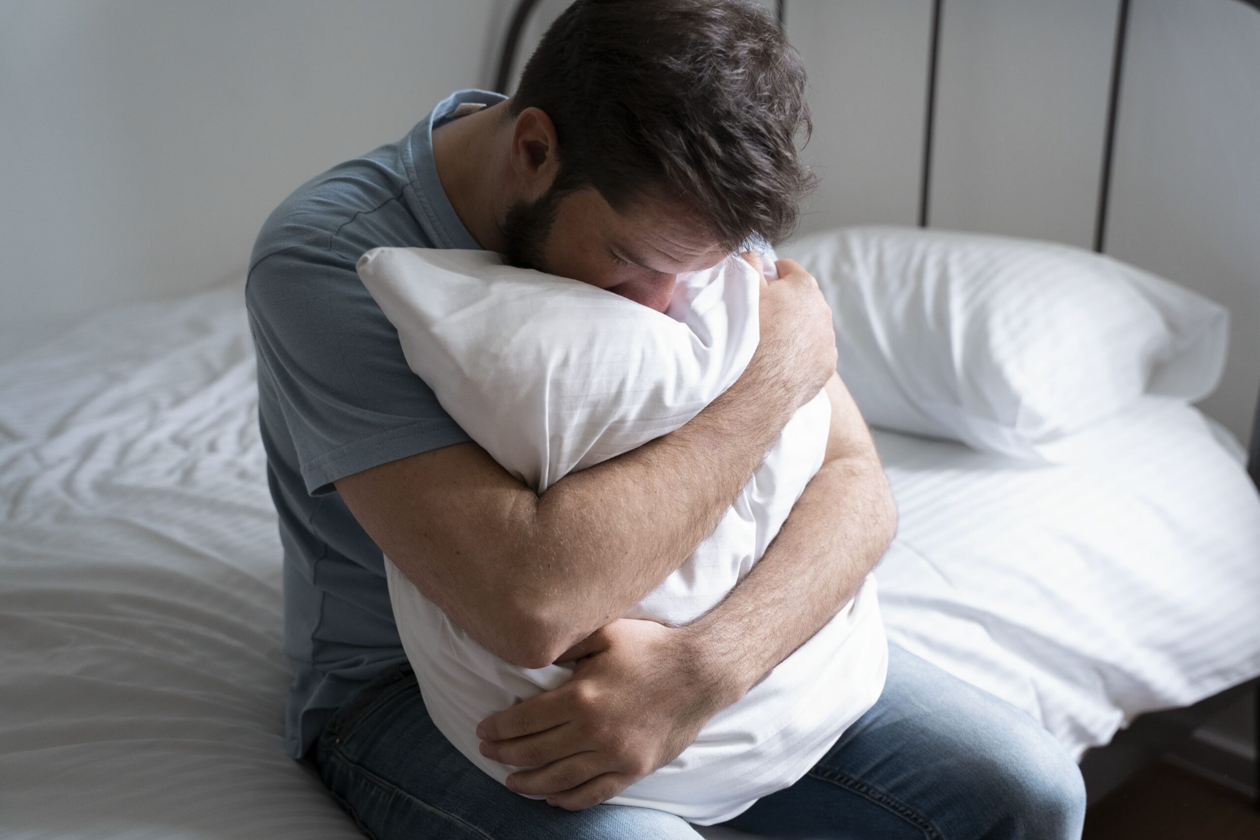 На фото зображений чоловік із темним волоссям та у сірій футболці. Він сидить на ліжку та обіймає обома руками подушку