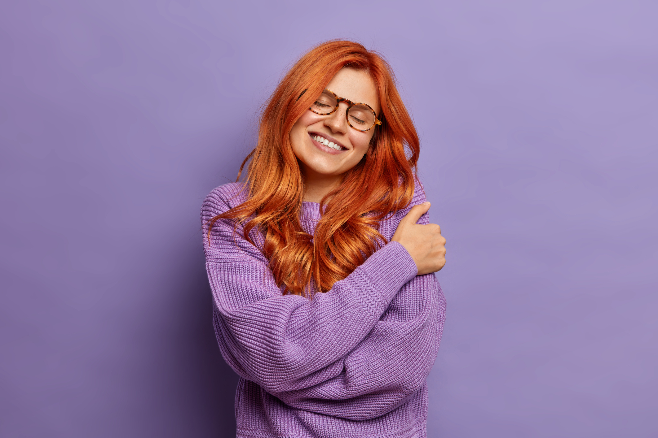 На фото руда жінка у бузковому светрі обіймає себе.