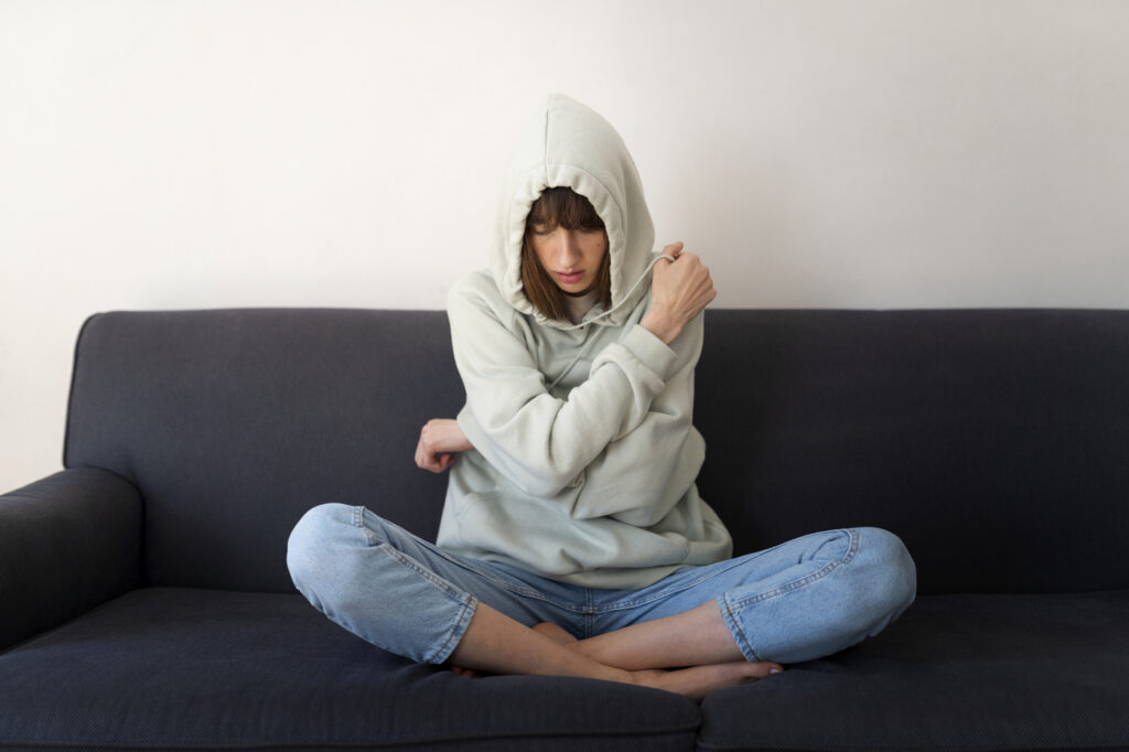 На фото зображена підлітка у світлих джинсах та худі, яка сидить на дивані. Вона натягнула капюшон на голову та обхопила себе руками.