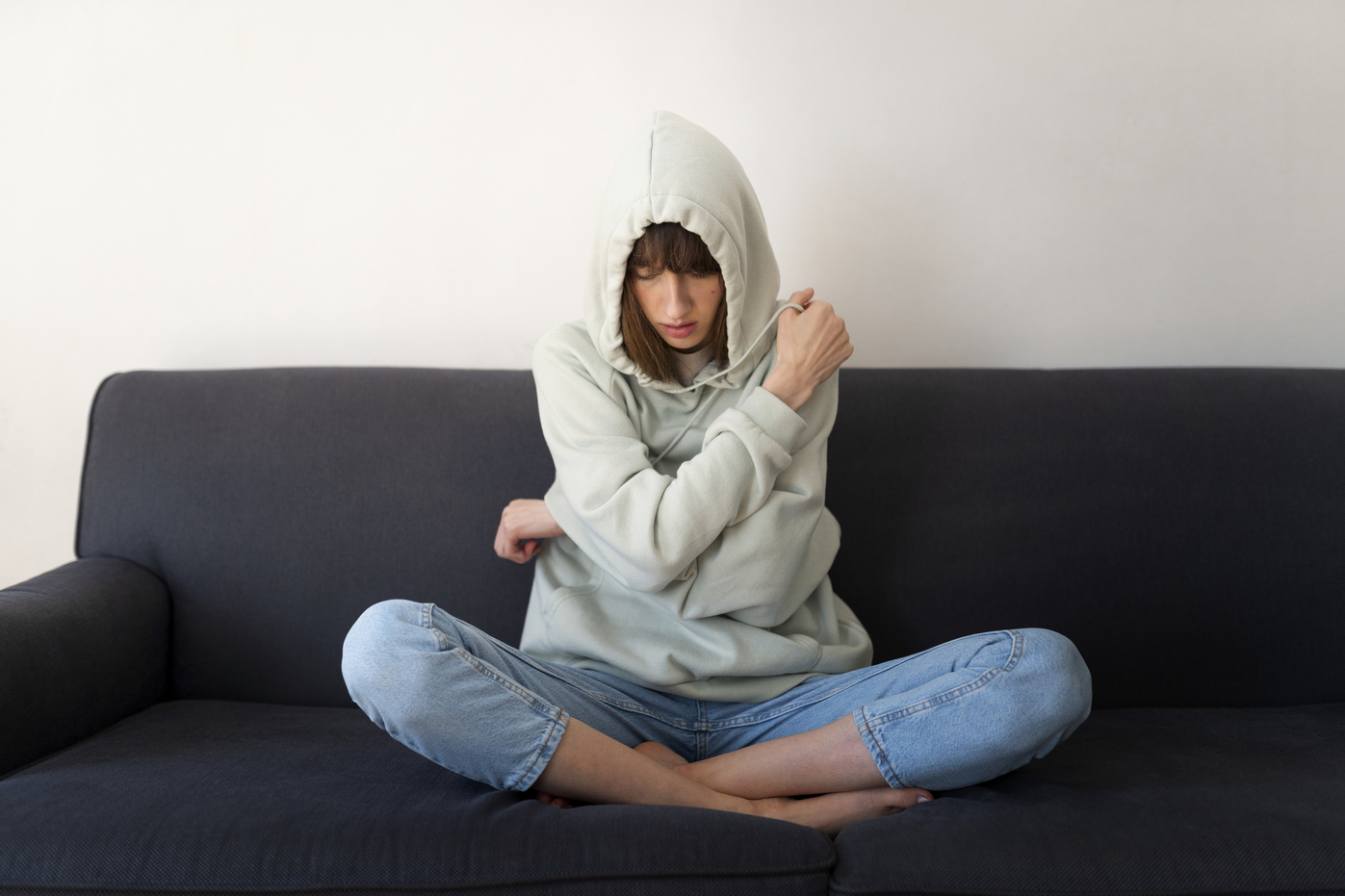 The photo shows a teenage girl wearing light jeans and a hoodie, sitting on a couch. She has pulled the hood over her head and wrapped her arms around herself