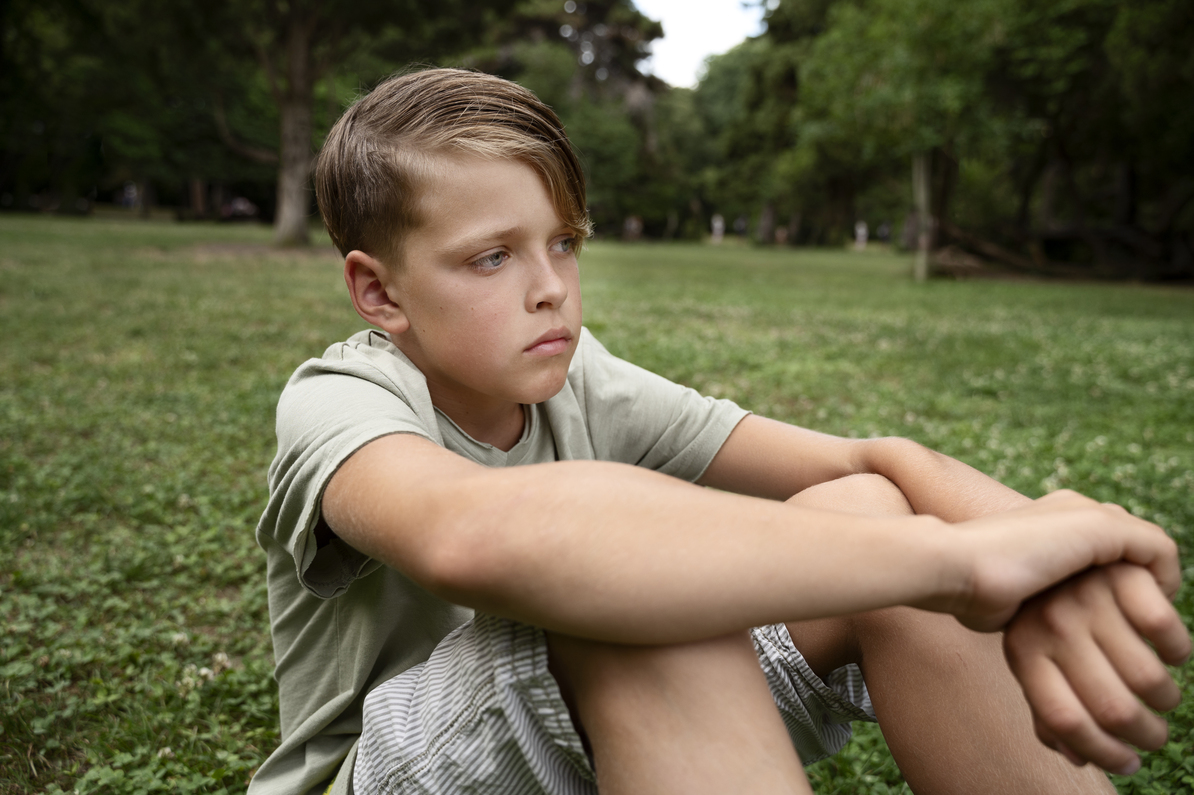 A boy is sitting on the grass in a park, hugging his knees, and looks upset and confused